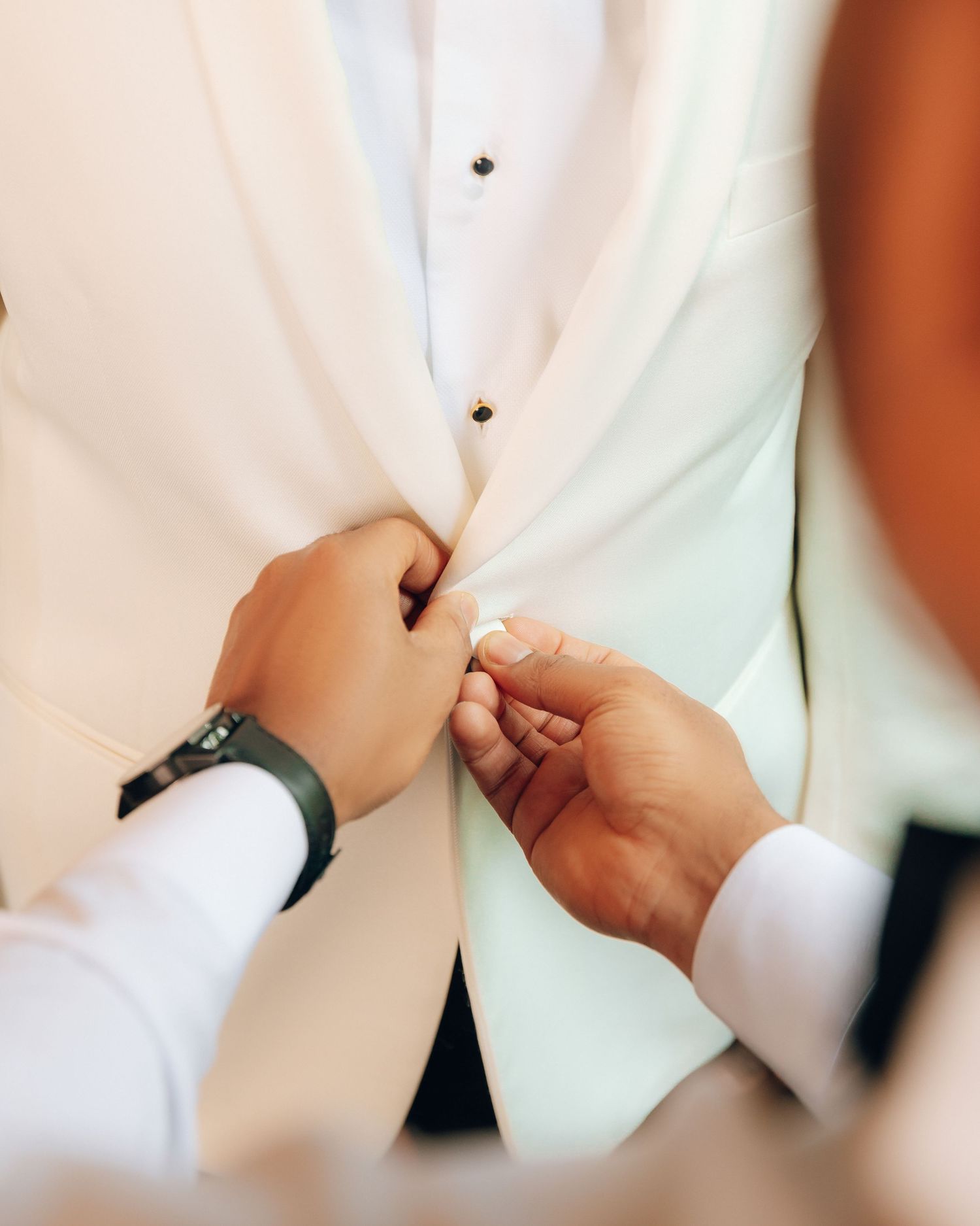 Hands help button up a white formal vest or waistcoat, showing preparation for a special event.