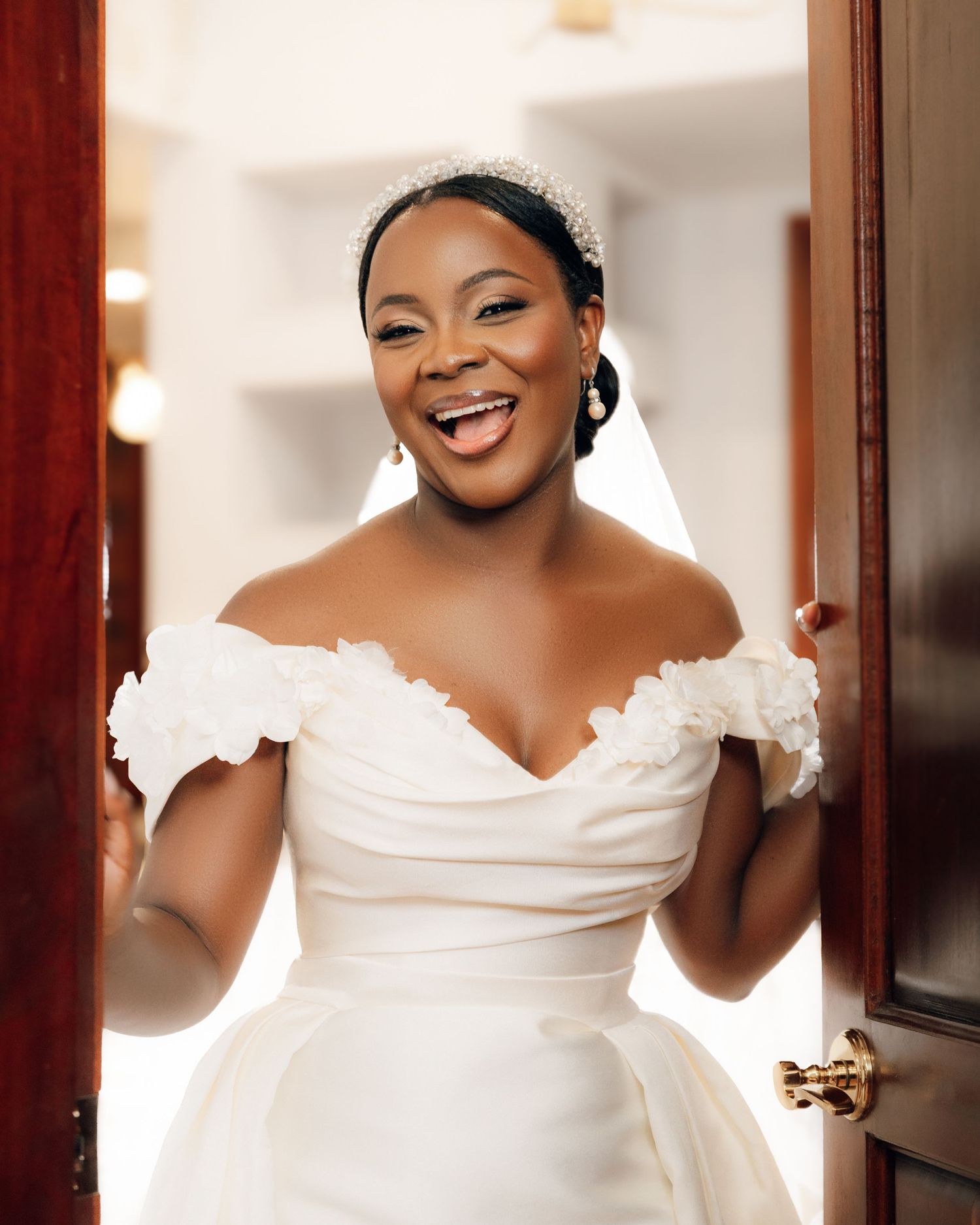 A radiant bride wearing an elegant off-shoulder white wedding gown stands by a doorway with a joyful expression.