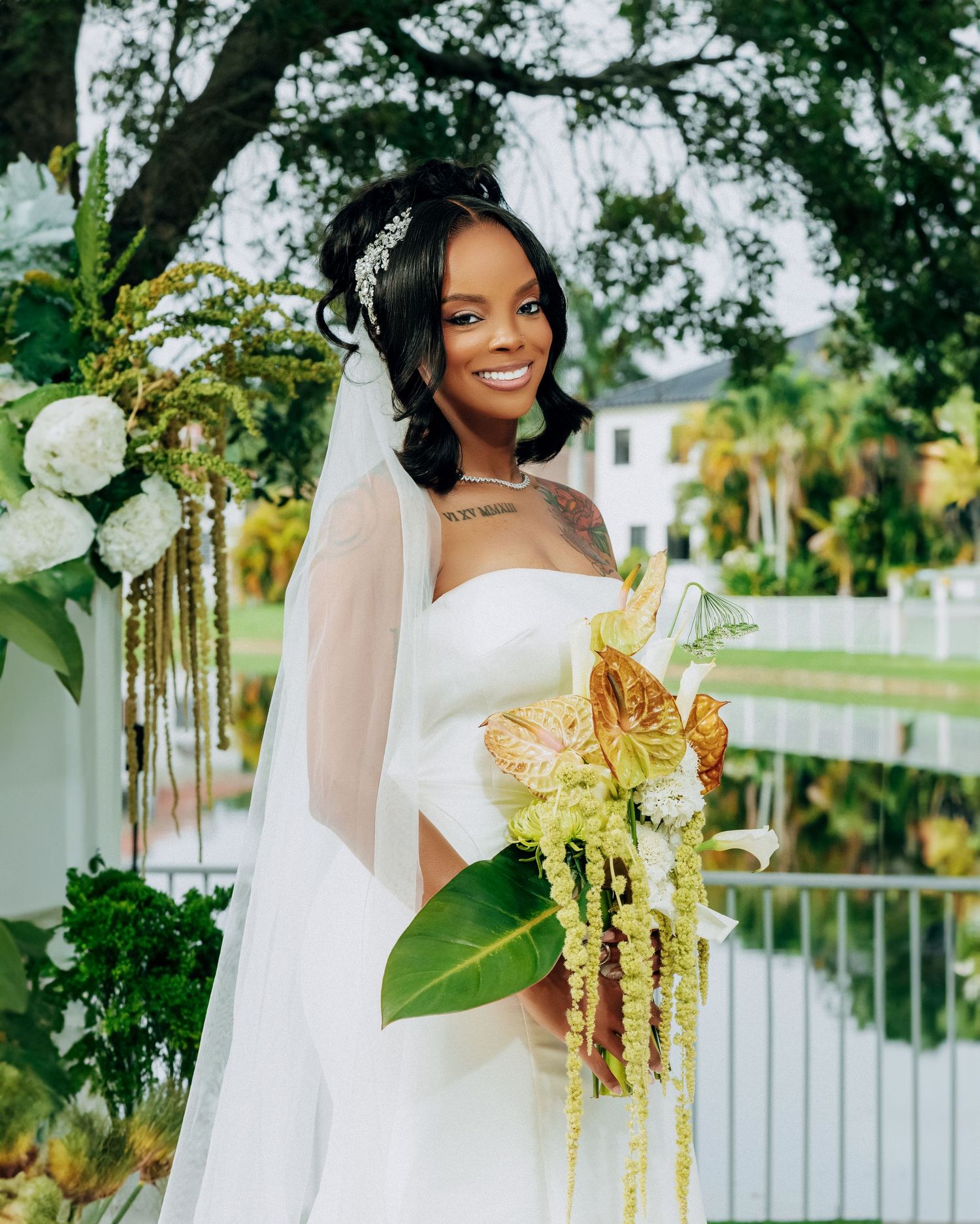 Bride in white gown holds unique gold and green tropical bouquet against garden backdrop.