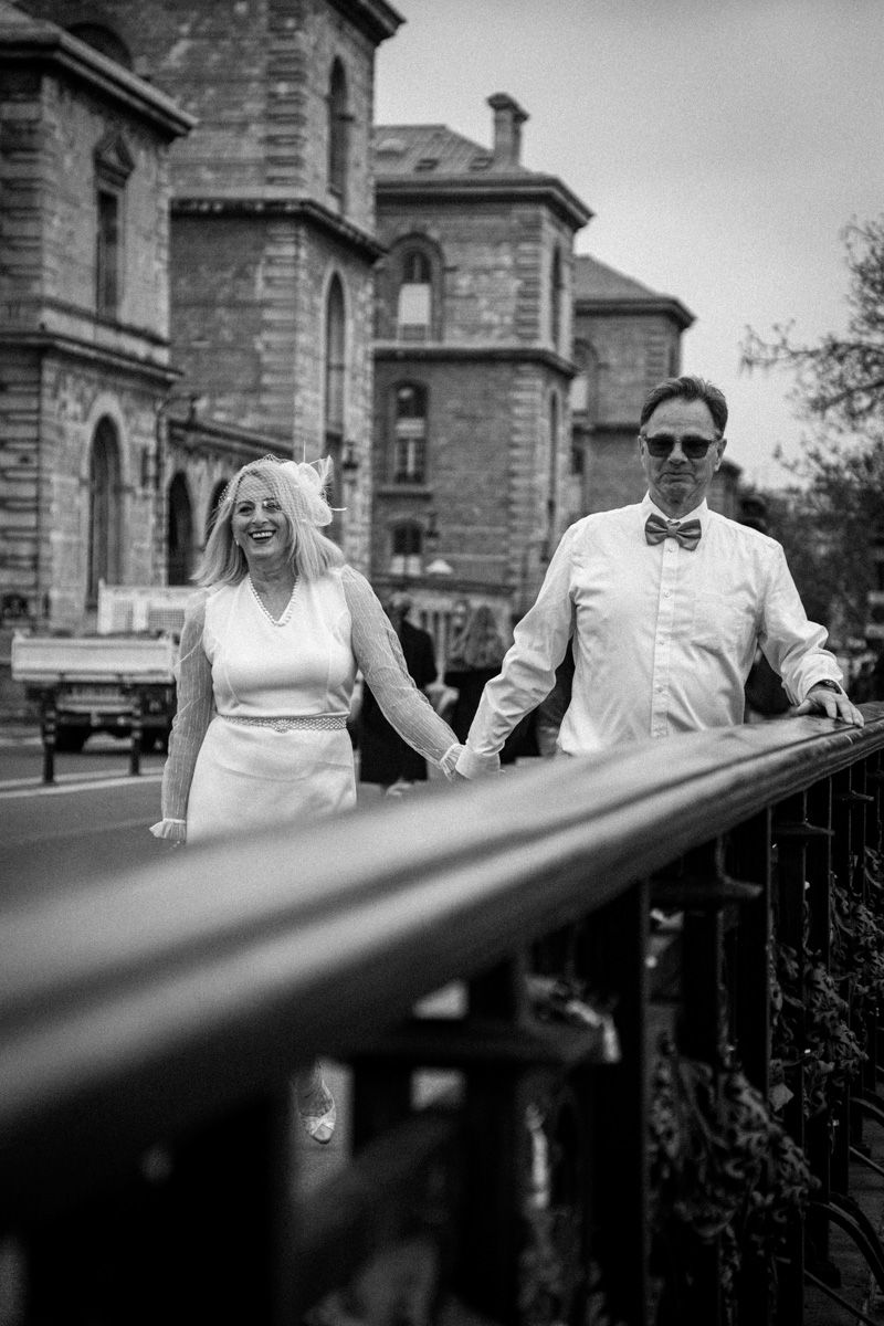 Black and white photo of a couple walking along a railing with historic stone building in background.