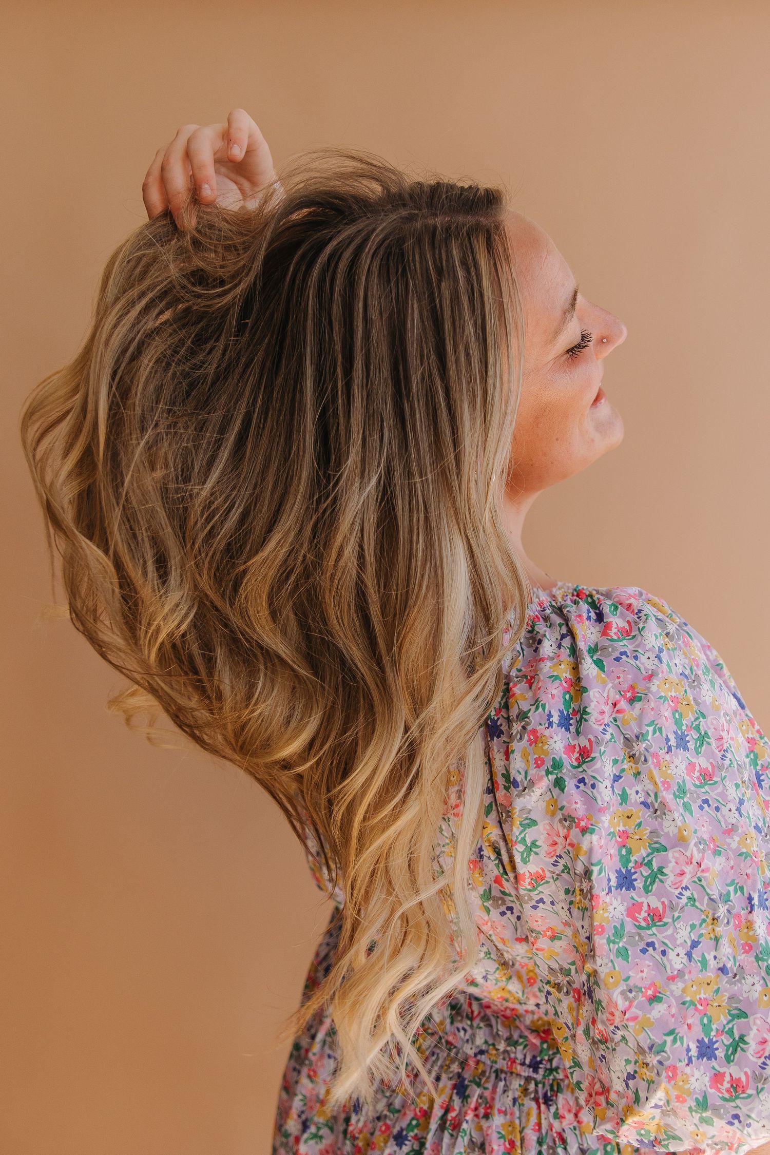 Side profile of wavy blonde hair and floral dress against peachy brown background, showing joyful movement and style.