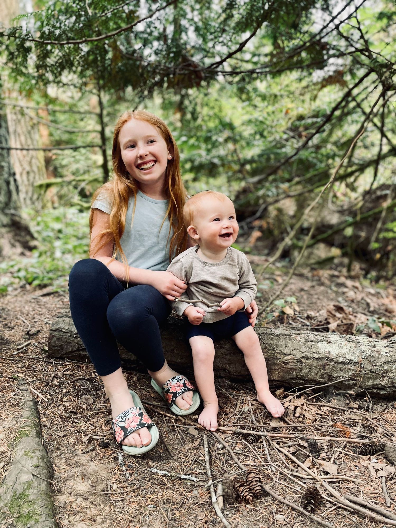 Siblings share a joyful moment sitting on tree roots in a lush forest setting.