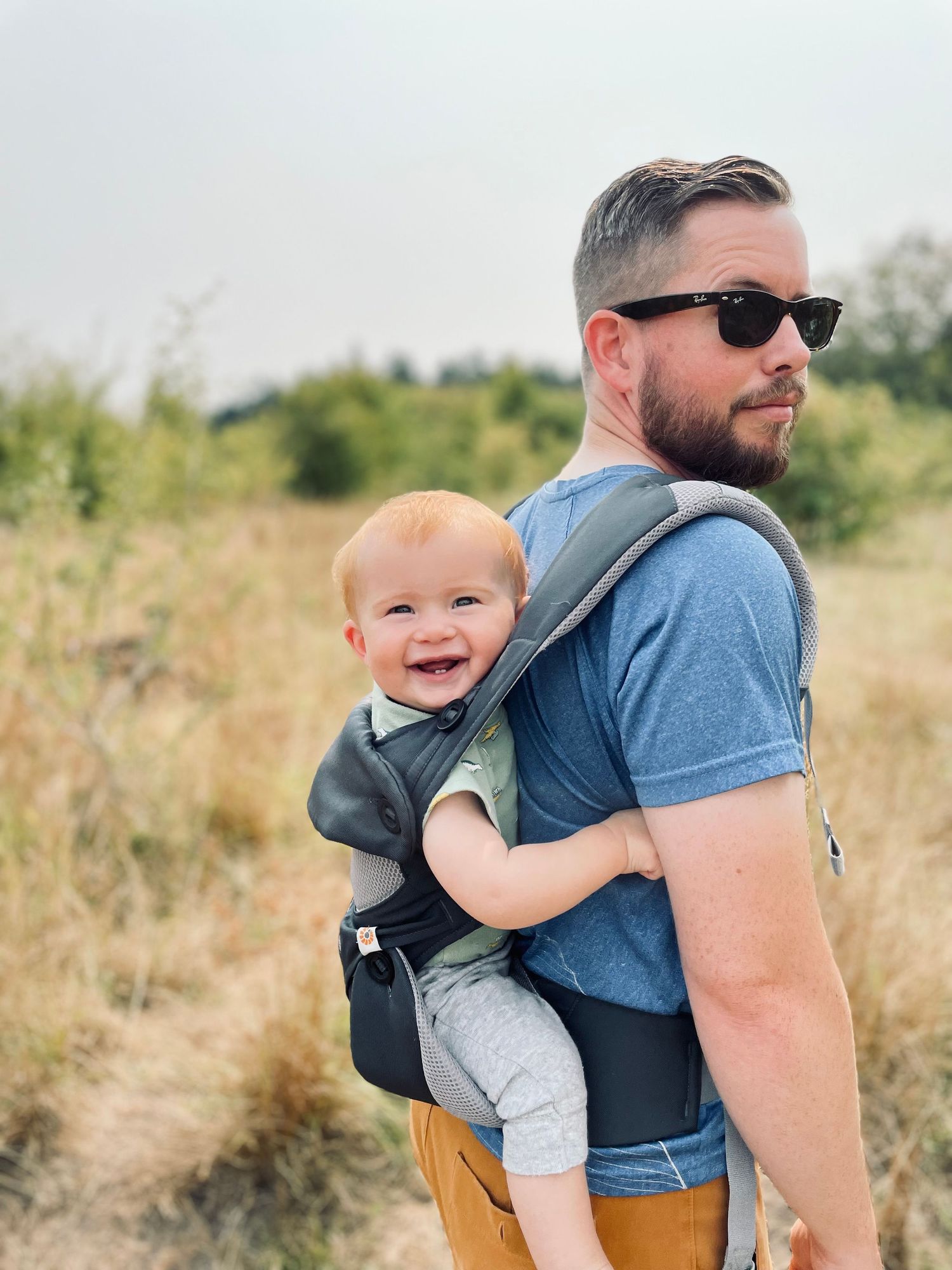 Baby smiles from carrier on parent's back during outdoor hike.