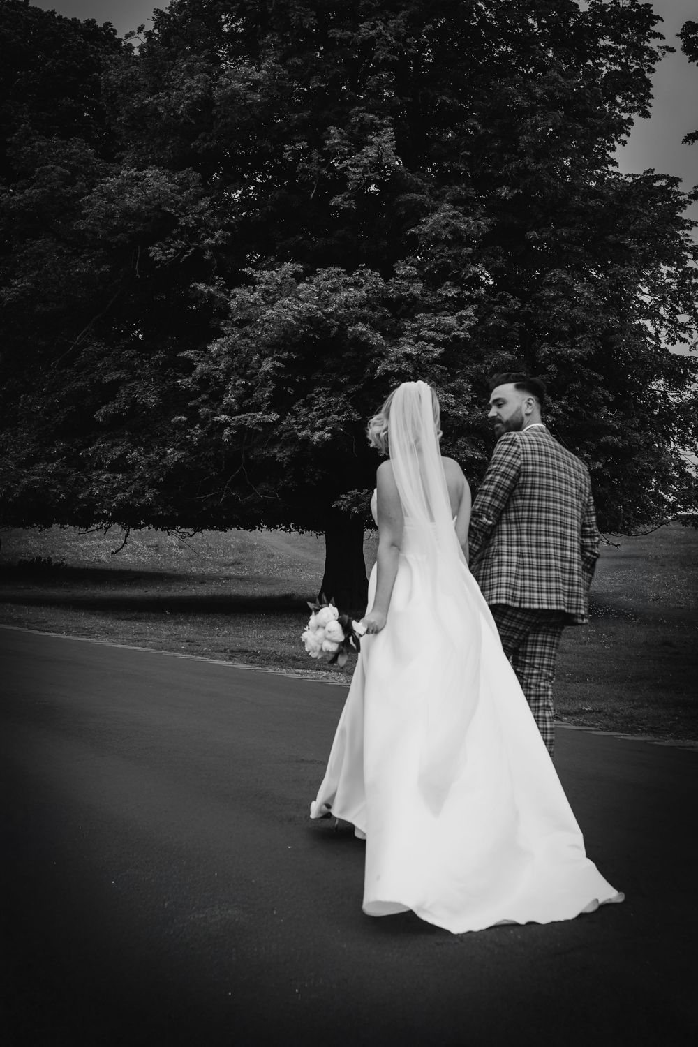 A black and white wedding photo capturing a bride in flowing white gown and groom in plaid suit walking on a path.
