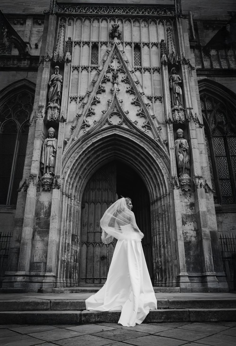 Dramatic black and white photo of a flowing white wedding dress against ornate Gothic church doorway architecture.