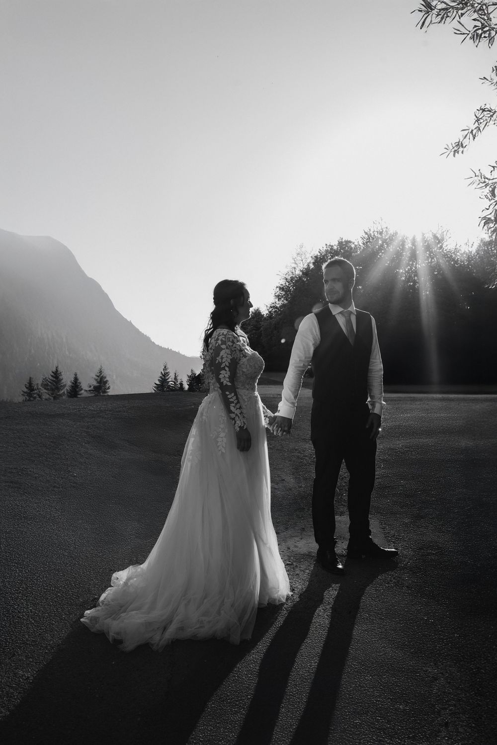 A dramatic black and white wedding photo of a couple holding hands at sunset with mountain silhouettes in the background.