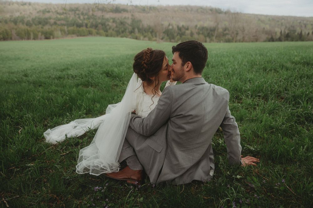A romantic wedding couple sits together in a lush green field.