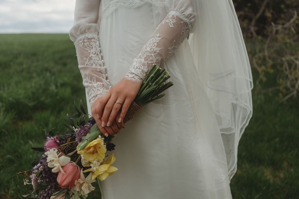 A close-up detail of lace sleeves and spring bouquet with pink and yellow flowers against a grassy field backdrop.