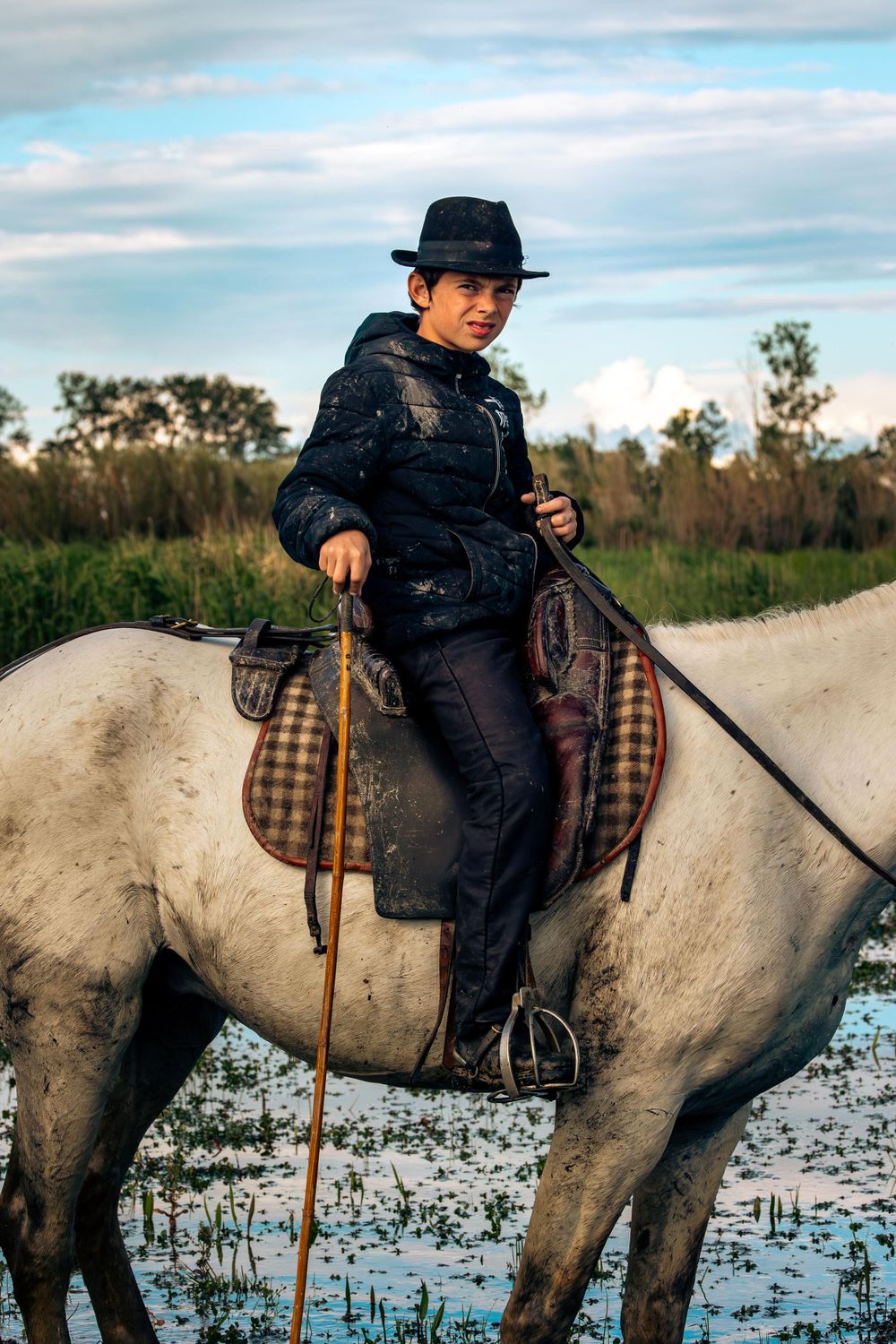 A rider in traditional garb sits atop a white horse in marshland.