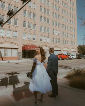 A couple in wedding attire stands outside a vintage-style brick apartment building with parked cars on a sunny day.