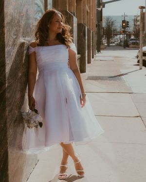 A person in a flowing white dress stands on a sunny urban sidewalk holding a small bouquet of flowers.