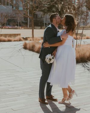 A couple shares a romantic kiss on their wedding day, standing on a paved pathway with winter foliage in the background.