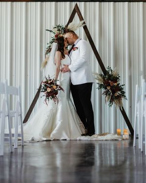 A couple shares a tender moment under a wooden triangular arch decorated with flowers at a modern indoor wedding ceremony.