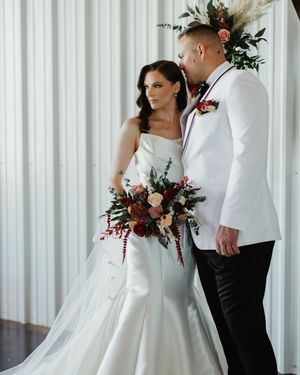 A couple in wedding attire poses against white curtains with burgundy and white florals and greenery.