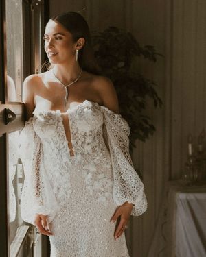 A bride in a vintage-inspired lace wedding dress with long puff sleeves stands elegantly by a window.