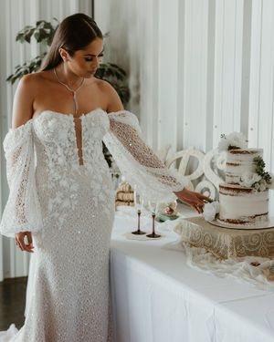Elegant off-shoulder lace wedding dress with dramatic bell sleeves shown next to a white tiered wedding cake.