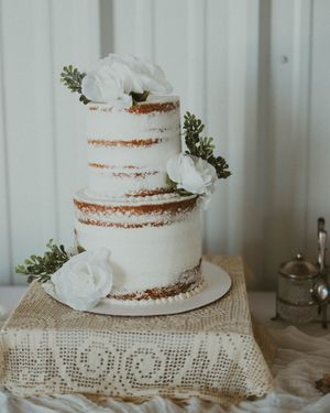 A rustic semi-naked wedding cake decorated with white roses and greenery on a lace tablecloth.