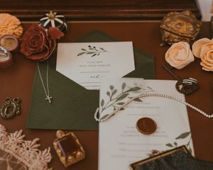 Elegant wedding invitation suite with green envelope, wax seal, and floral accents laid on wooden surface with vintage decor.