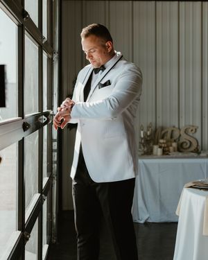 A groom in a white tuxedo jacket adjusts his bow tie while getting ready next to a window.
