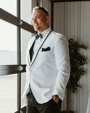 A groom in a white dinner jacket with black bow tie and pocket square smiles while standing by a window.