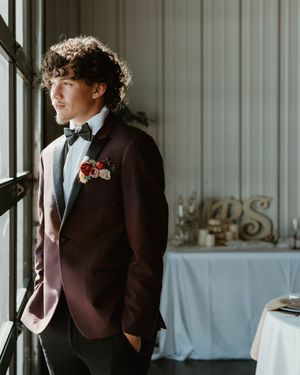 A young person in a burgundy tuxedo jacket and bow tie stands by a window at a formal event.