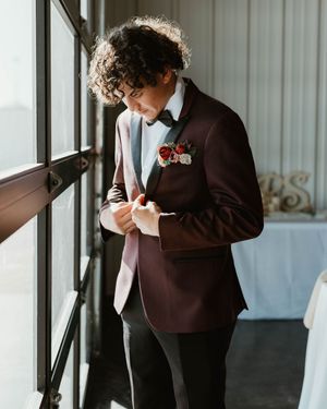 A groom in a burgundy suit adjusts his tie while standing by a window.