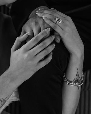 Black and white close-up of hands touching with jewelry, bracelets and small tattoo visible.