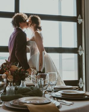 A romantic wedding couple shares an intimate moment by a window with an elegant table setting in the foreground.