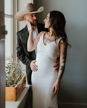 A romantic couple in country western attire share an intimate moment by a window with a potted plant.