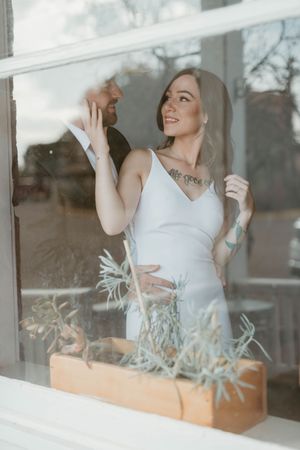 Woman in white dress smiling while standing by window with a planter box during a photoshoot.