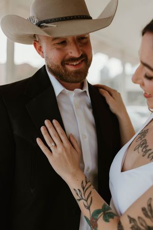 Close up photo of couple with cowboy hat and white dress showing wedding ring and tattoos.