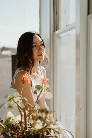 A dreamy portrait by a window with soft lighting and coral-colored flowers in the foreground.