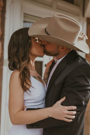 Romantic couple embraces in soft golden light, with a tan cowboy hat and formal attire creating a rustic elegant mood.