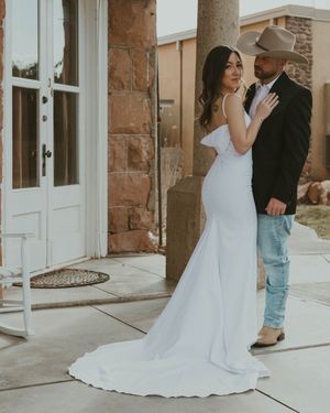 Elegant bride in fitted white gown poses with partner in cowboy hat on stone porch of rustic building.