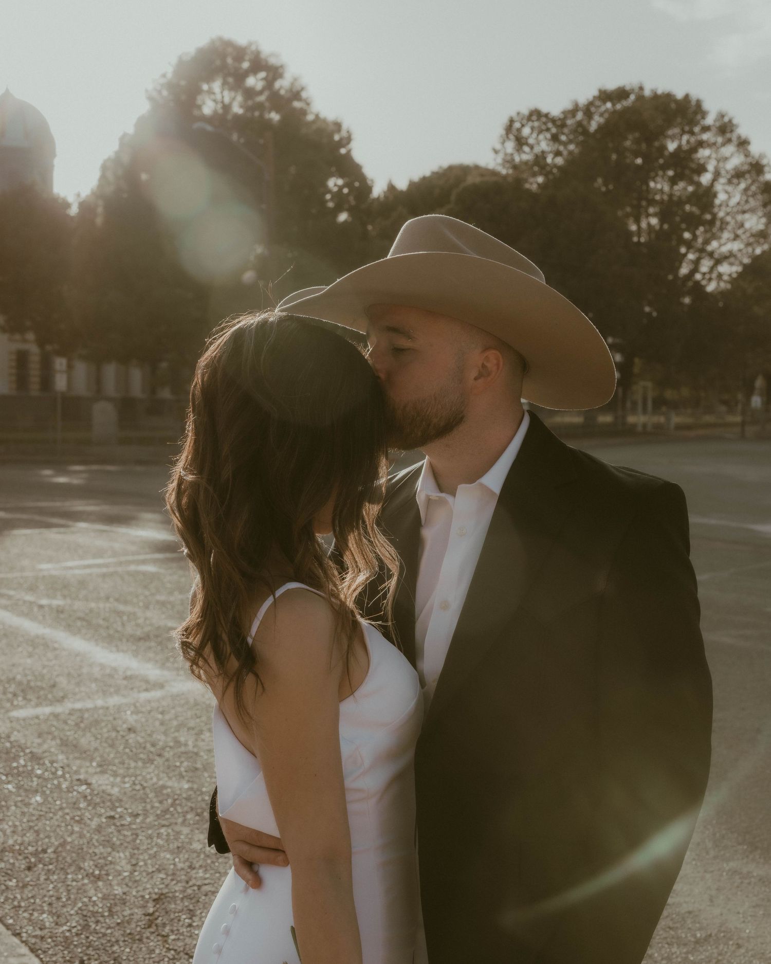 Romantic sunset portrait of newlyweds embracing with cowboy hat silhouetted against evening sky.