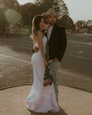 Couple in wedding attire poses together at sunset in a rustic outdoor setting with the bride holding a bouquet.