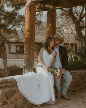 Couple in wedding attire sits together under rustic wooden gazebo with stone base in outdoor setting.