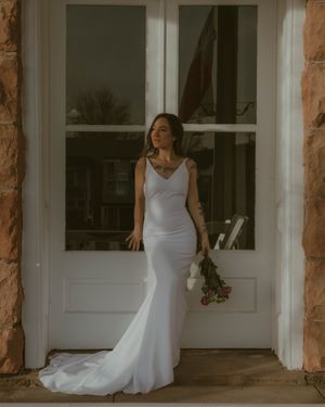 A figure in an elegant white satin wedding dress stands in a doorway against stone columns in soft natural light.