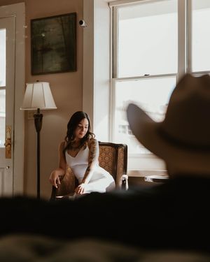 A person sits in a vintage armchair near bright windows in a room with warm lighting and minimalist decor.