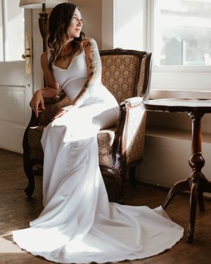 Elegant white wedding gown with fitted silhouette and flowing train drapes beautifully over vintage upholstered armchair.