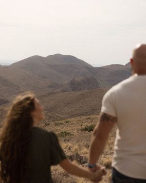 A couple holding hands while overlooking a scenic desert mountain landscape at sunset.