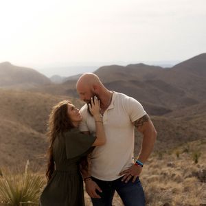 A romantic couple poses in the desert against a backdrop of rugged mountains and desert brush in golden light.