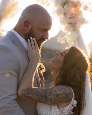 A tender romantic moment between newlyweds in formal wedding attire against a soft floral backdrop outdoors at sunset.