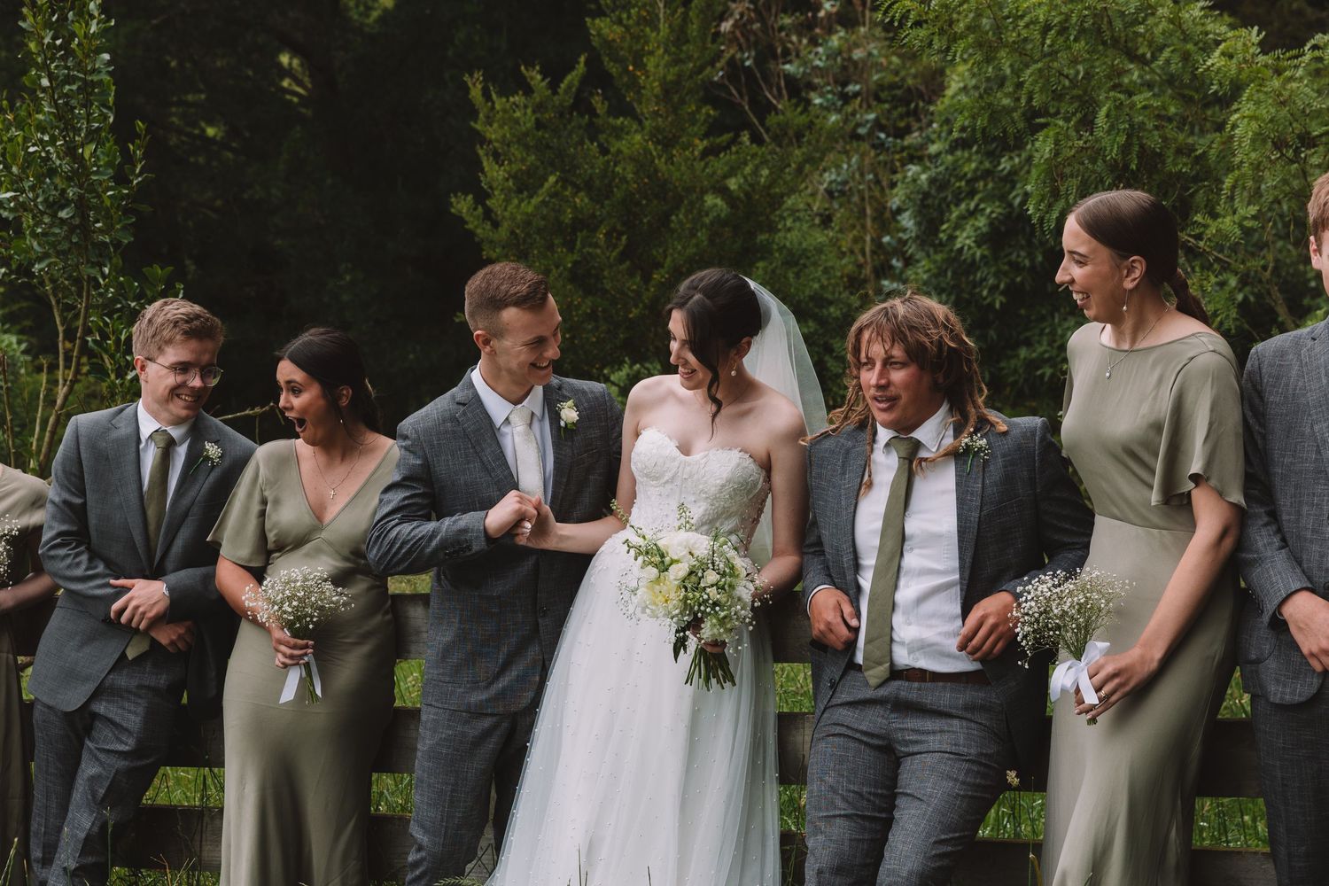 Wedding party shares candid moments while lined up against a wooden fence in an outdoor garden setting.