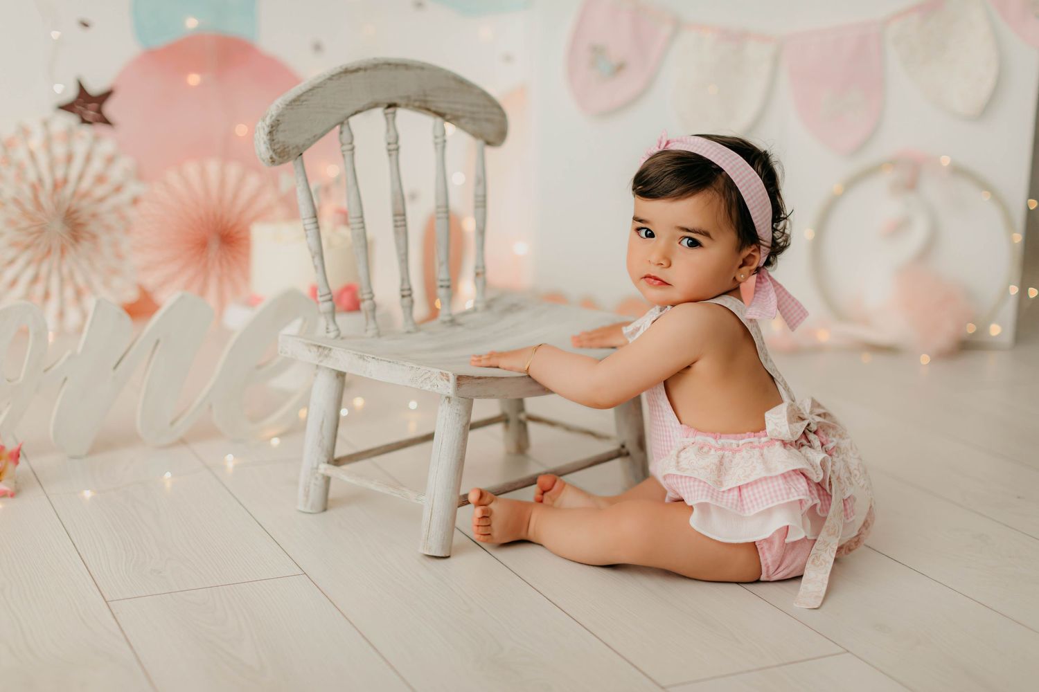 Fotografía del primer añito de una niña con una cinta de pelo y peto vichy rosa sentada con decoración en tonos rosas.
