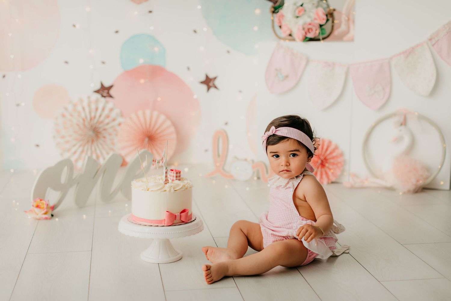 Fotografía del primer añito de una niña con una cinta de pelo y peto vichy rosa con decoración en tonos rosas.