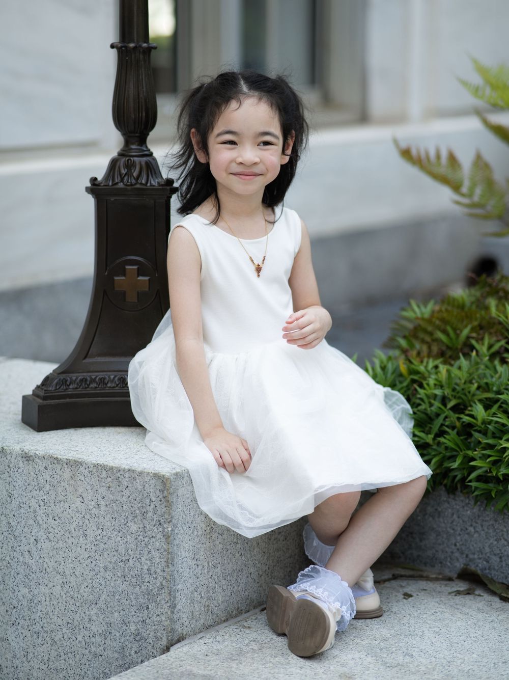 A young child in a white sleeveless dress sits casually on a concrete ledge near an ornate lamppost on a sunny day.