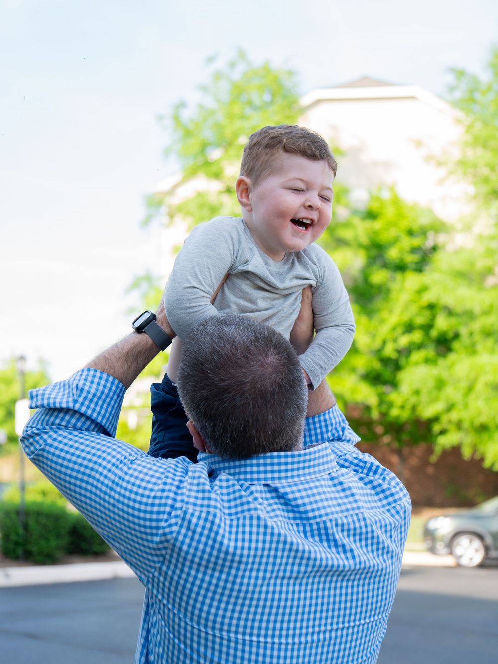 A person in a blue checkered shirt lifts and spins a child wearing grey outdoors against green foliage.