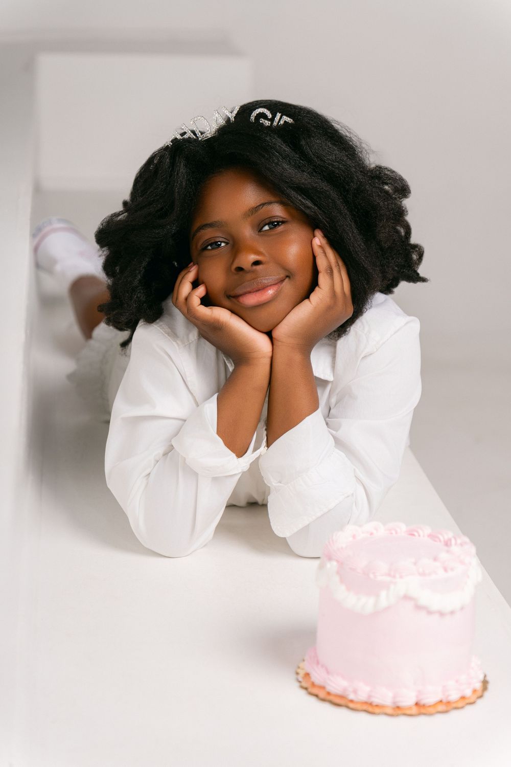 A person in a white shirt rests their head in their hands while smiling next to a pink frosted cake.
