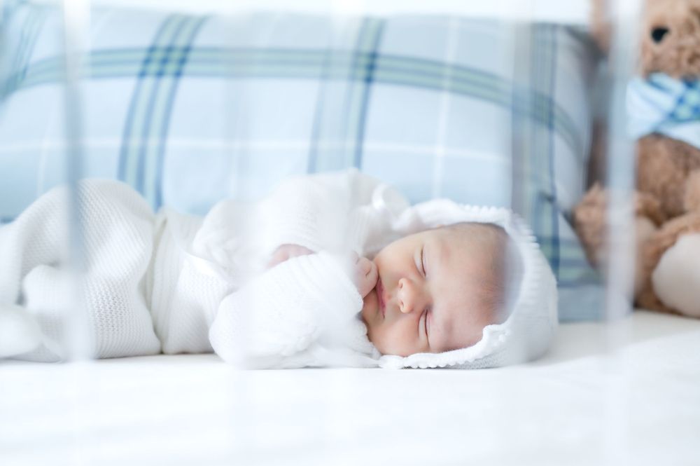 Peaceful newborn baby sleeping on plaid bed with teddy bear visible in background.